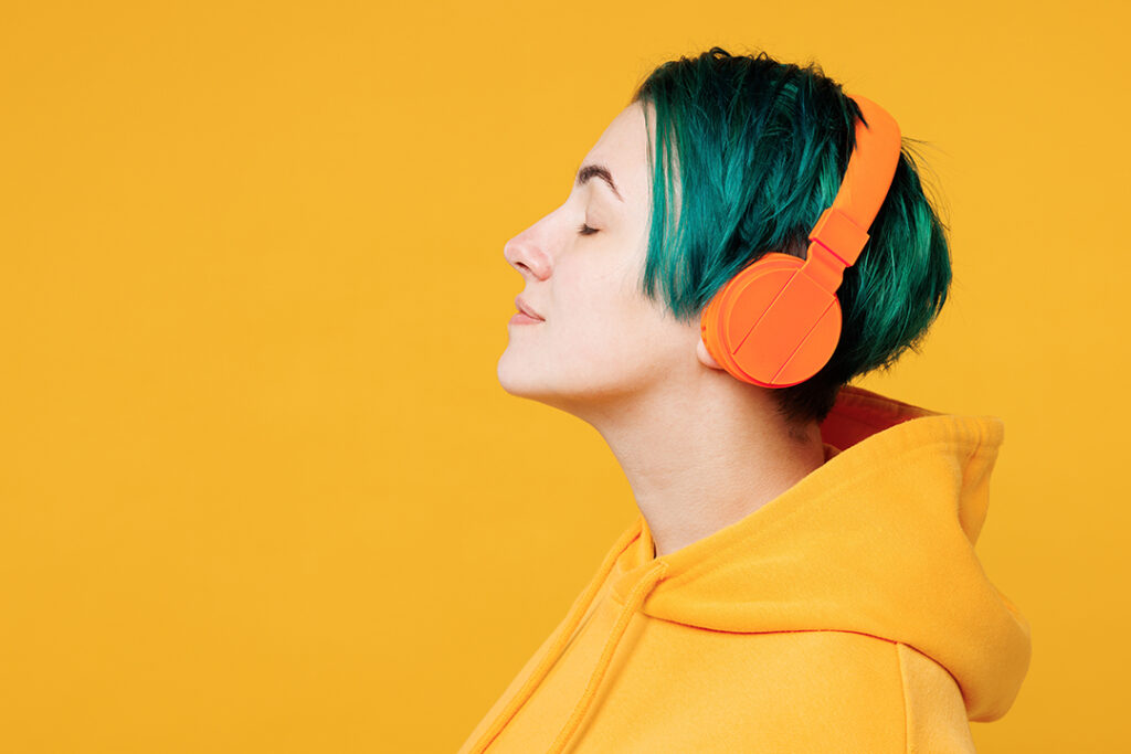 Närbild av ung rolig glad kvinna med färgat grönt hår i hoodie, casual kläder, lyssnar på musik i hörlurar isolerad på vanlig gul orange bakgrund,