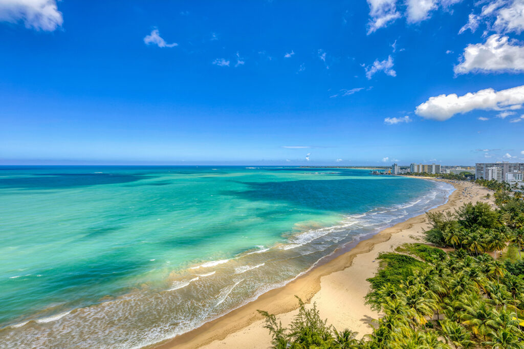 
Flygfoto över Isla Verde-stranden i Carolina, Puerto Rico, med vidsträckt gyllene sand, klart turkost vatten och en bakgrund av palmer och kustnära bostadshus under en blå himmel.