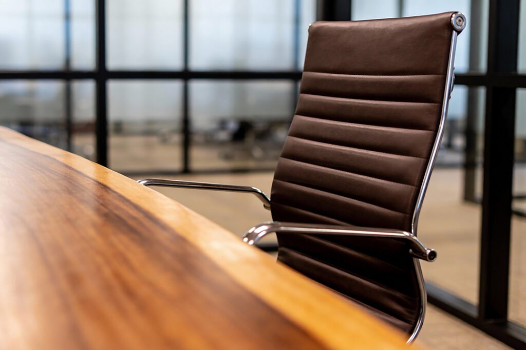 En ensam chefsstol med brun läderklädsel står bredvid ett glänsande konferensbord i trä, med en suddig kontorsinredning synlig genom glasväggar.
