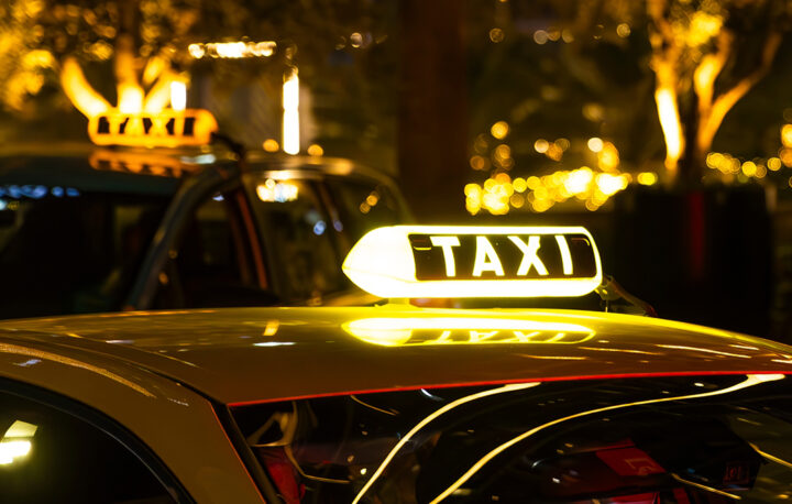 En gul taxi med en upplyst TAXI-skylt på taket. Taxinen är parkerad i en stad på natten, med suddiga ljus i bakgrunden.